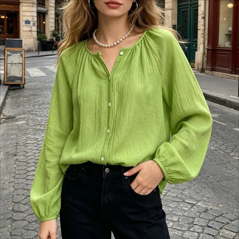 A woman wearing a light lime green long-sleeve blouse with a gathered round neckline and front buttons, paired with black jeans, standing on a cobblestone street.