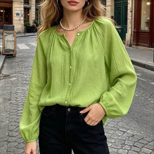 A woman wearing a light lime green long-sleeve blouse with a gathered round neckline and front buttons, paired with black jeans, standing on a cobblestone street.