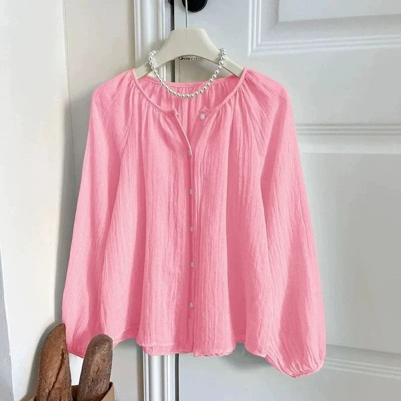 A pink long-sleeve button-up blouse with a gathered round neckline, hanging on a white hanger decorated with a pearl necklace, set against a white paneled door.