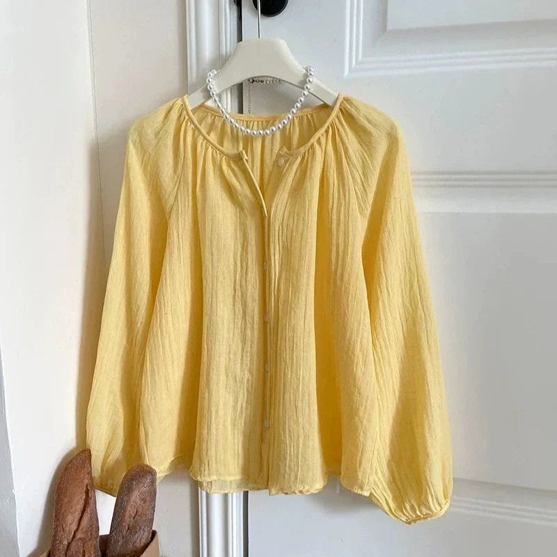 A pale yellow crinkle-textured blouse with long balloon sleeves and a gathered round neckline, hanging on a white hanger against a white paneled door. A pearl necklace is draped around the collar area.