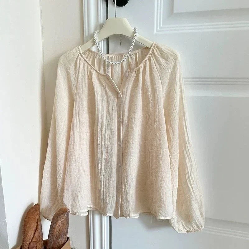 A light beige crinkled long-sleeve blouse hanging on a white hanger with a pearl necklace, set against a white paneled door background.