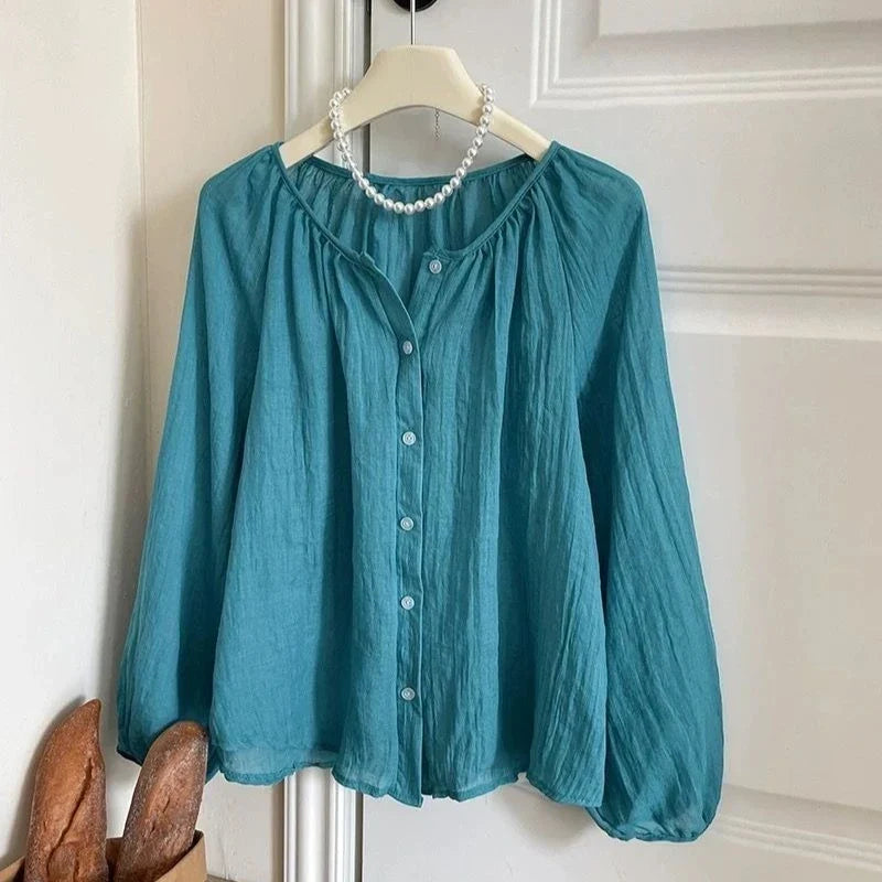 A teal blue crinkled long-sleeve blouse hanging on a white hanger with a pearl necklace, set against a white paneled door background.