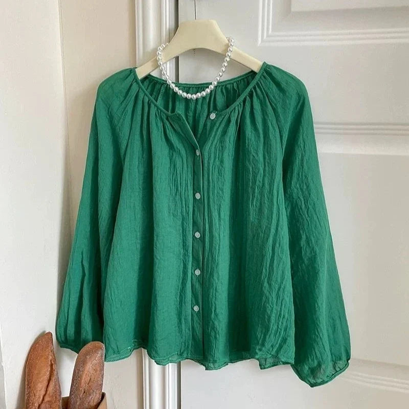 A green long-sleeve blouse with a gathered round neckline and front button closure, hanging on a cream hanger against a white paneled door. A pearl necklace is draped around the neck area.