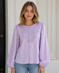 A woman wearing a lavender long-sleeve blouse with floral embroidery and a button-up front, paired with blue jeans, standing in front of a white paneled door.