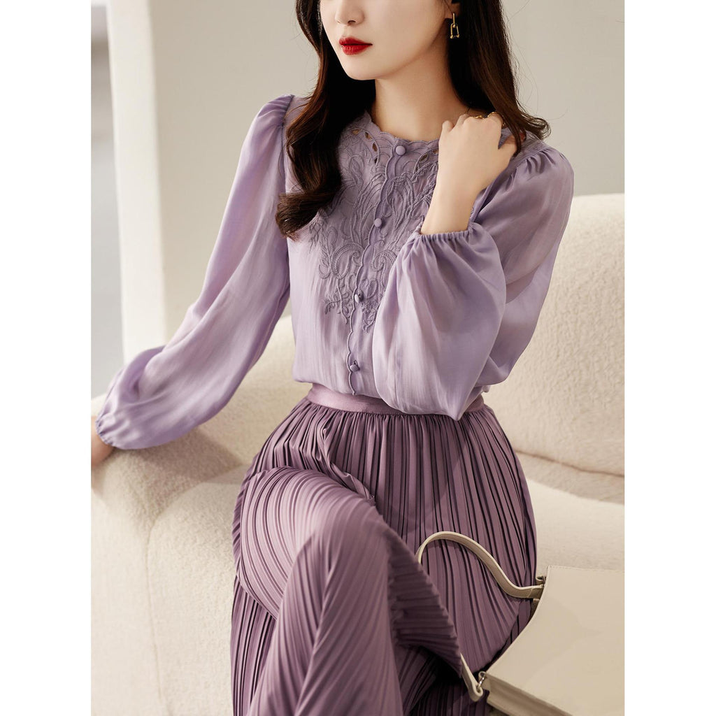 A woman seated on a cream sofa wearing a lavender two-piece outfit consisting of a long-sleeve embroidered blouse and matching pleated skirt, with a metallic handbag beside her.