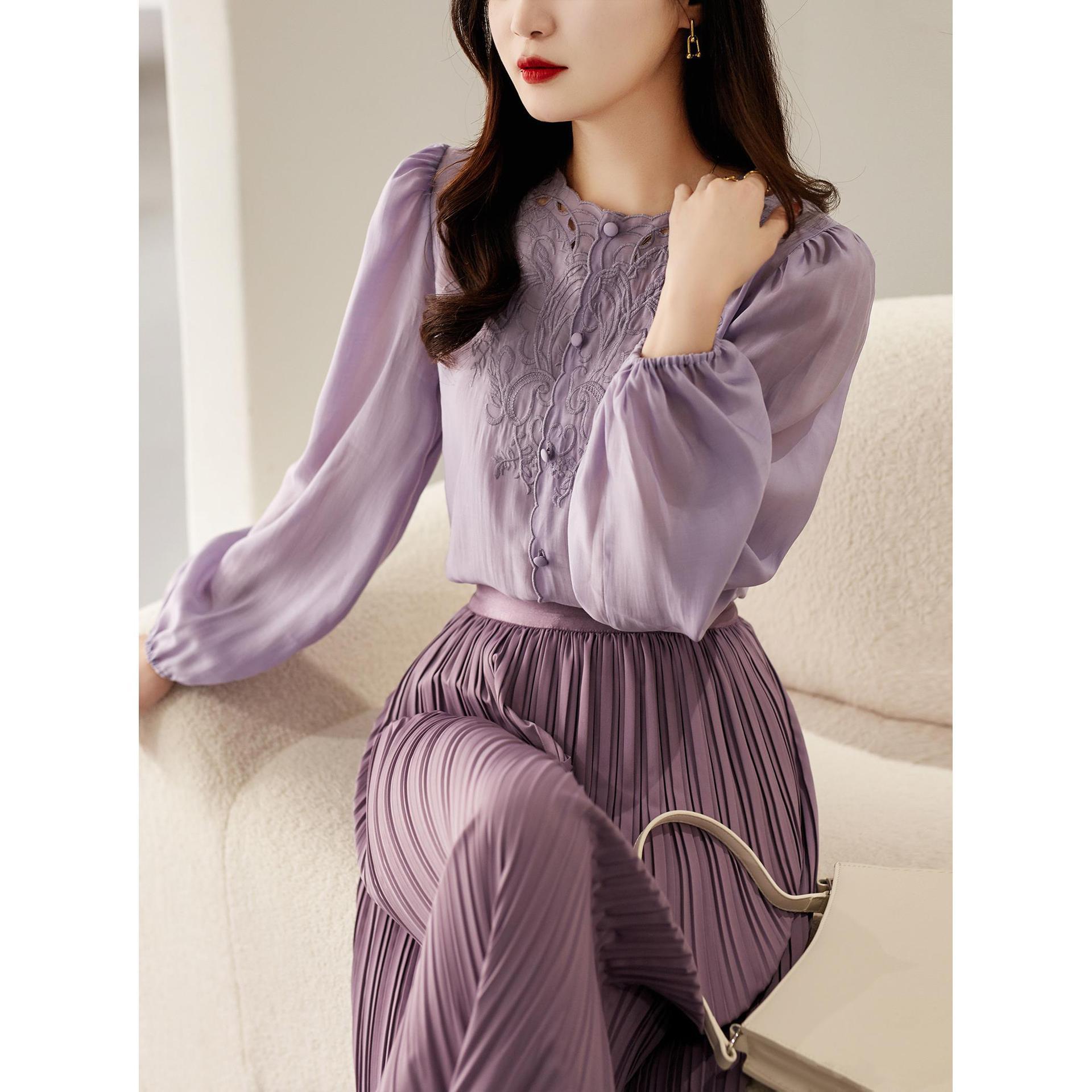 A woman seated on a cream sofa wearing a lavender two-piece outfit consisting of a long-sleeve embroidered blouse and matching pleated skirt, with a metallic handbag beside her.