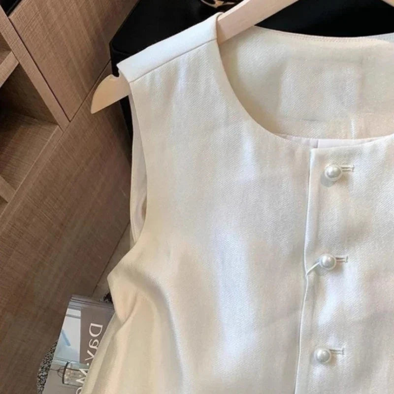 A cream-colored sleeveless vest with pearl-like buttons, hanging on a wooden hanger against a wooden shelving background.
