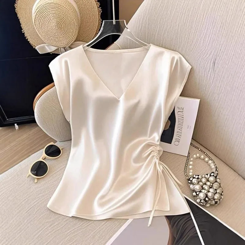 A cream-colored satin top with a V-neckline and side drawstring, styled on a hanger in a flat-lay arrangement with a straw hat, sunglasses, and a pearl bag.