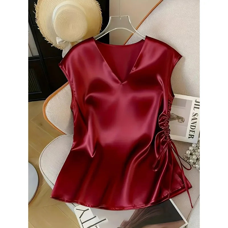 A sleeveless deep red satin top with a V-neckline and side ruching, hanging on a clear hanger on a chair.